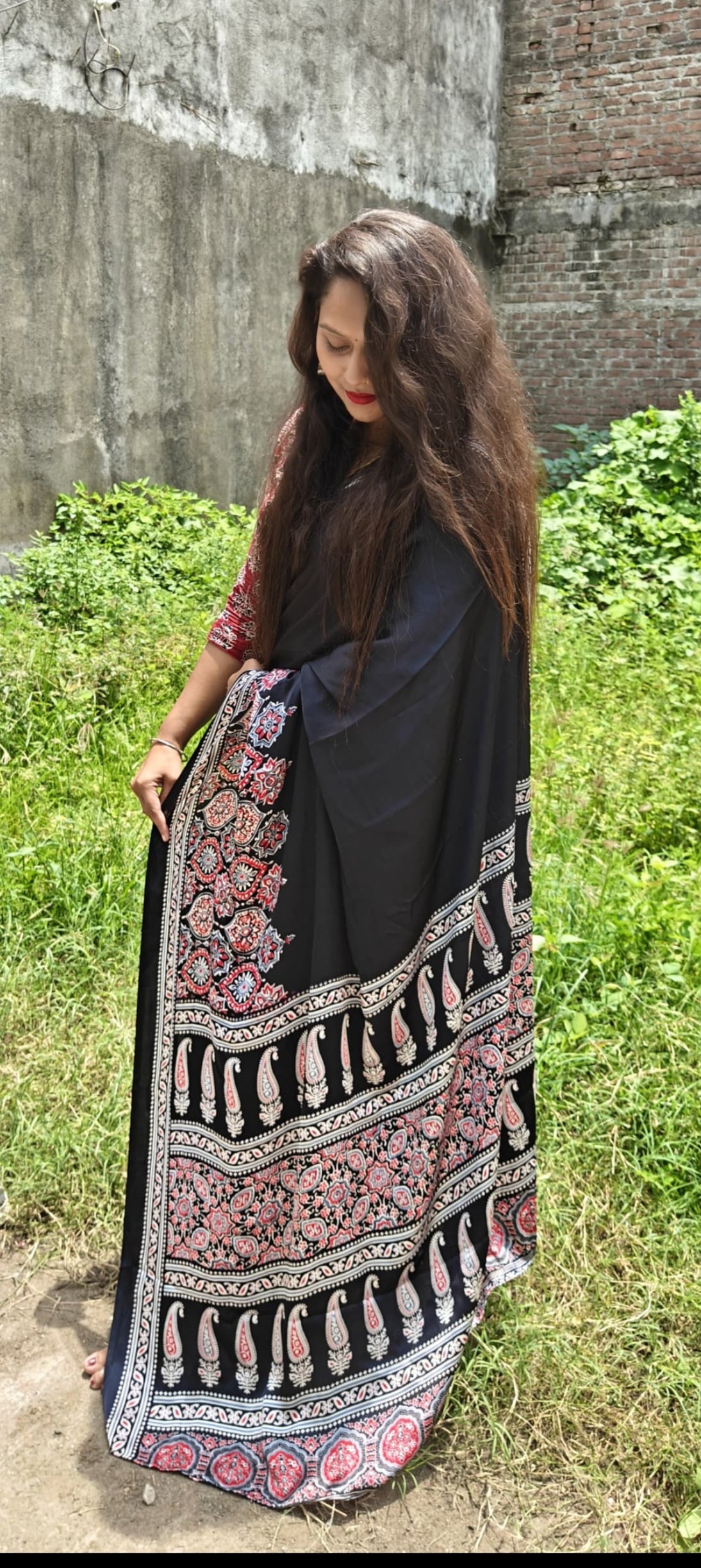 Woman wearing a black and patterned saree standing in front of a brick wall.