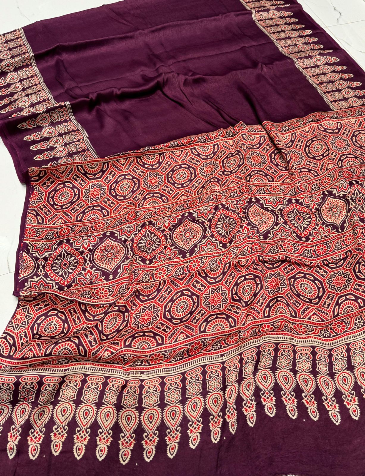 A wine-colored saree with traditional Ajrakh patterns, including geometric and floral designs, alongside a running blouse piece.